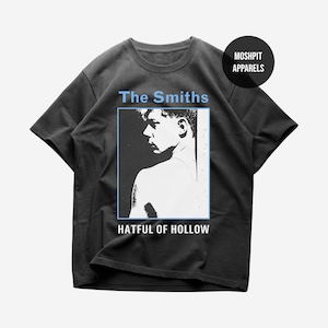 Op de afbeelding: Donkergrijs t-shirt met een zwart-wit afbeelding van een persoon. De tekst "The Smiths" staat in lichtblauw boven de afbeelding en "Hatful of Hollow" eronder. Het shirt heeft ook een "Moshpit Apparels" logo.