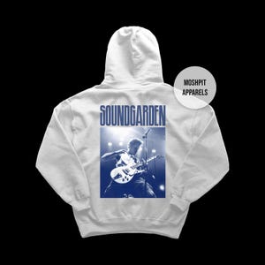 Puede incluir: Una sudadera con capucha gris claro con una imagen azul y blanca de un guitarrista. La palabra "SOUNDGARDEN" está impresa encima de la imagen. La sudadera con capucha tiene una capucha y un bolsillo delantero. La marca "MOSHPIT APPARELS" está impresa en el lado derecho.