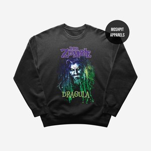 May include: Black crew neck sweatshirt featuring a graphic of Rob Zombie with the text "Rob Zombie Dragula" in purple and green. The image has a distressed, grunge style. The sweatshirt has a round neckline and long sleeves.