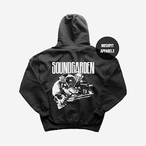 Puede incluir: Sudadera con capucha negra con el nombre de la banda "SOUNDGARDEN" en letras blancas. Debajo, un gráfico en blanco y negro de una calavera y un motor de motocicleta. La sudadera tiene capucha y un logo "MOSHPIT APPARELS".