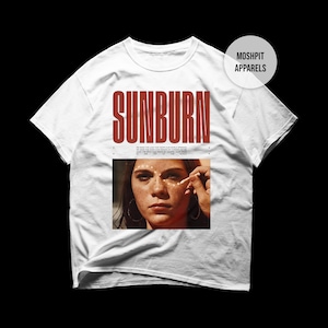 Pode incluir: Camiseta branca com a palavra "SUNBURN" em letras vermelhas grandes. Abaixo, uma foto de uma pessoa com sardas e o texto "MOSHPIT APPARELS" em um círculo branco no canto superior direito.