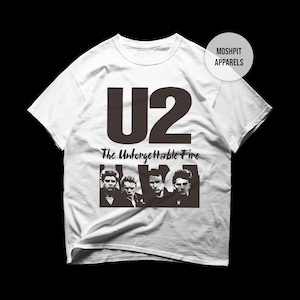 Può includere: T-shirt bianca con il logo degli U2 e la scritta "The Unforgettable Fire" in nero. Sotto il testo c'è un'immagine in bianco e nero dei membri della band. La maglietta ha un girocollo e maniche corte.