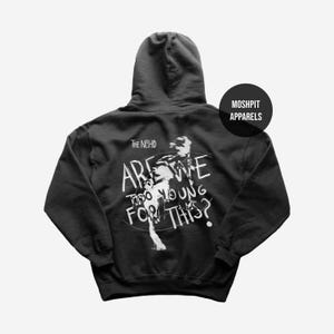 May include: Dark gray hoodie with the text "ARE WE TOO YOUNG FOR THIS?" in white, along with a graphic of a person. The band name "The NBHD" is above the text. A black circle with "MOSHPIT APPARELS" is on the right.