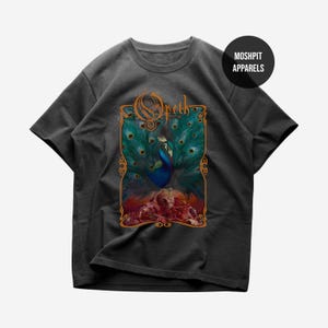 Puede incluir: Camiseta gris oscuro con un diseño de pavo real con plumas verde azulado y azules, sobre un fondo colorido. La camiseta incluye el nombre de la banda "Opeth" en una fuente naranja adornada. El nombre de la marca "Moshpit Apparels" está en un círculo negro.