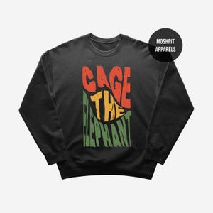 May include: Black crewneck sweatshirt with the text "CAGE THE ELEPHANT" in red, yellow, and green. The text is stylized with a leaf design. The brand name "MOSHPIT APPARELS" is in a black circle in the upper right corner.