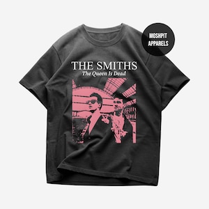 Op de afbeelding: Donkergrijs T-shirt met een roze afbeelding van twee mannen en de tekst "THE SMITHS The Queen Is Dead". Het shirt heeft een ronde hals en korte mouwen. Een rond logo met de tekst "MOSHPIT APPARELS" staat rechtsboven.