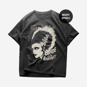 May include: Dark gray t-shirt featuring a vintage-style graphic of a woman with dramatic makeup and the text "The Bride and the Banshees." The shirt also includes the text "Following the footsteps of a rag doll dance we are entranced."