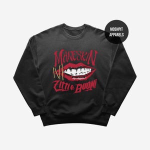 May include: Black crewneck sweatshirt with a red graphic of a mouth with the word "MANESKIN" above it and "Zitti E Buoni" below. The graphic also includes the letter "N" and two gold-colored bars. A black circle with white text that says "MOSHPIT APPARELS" is in the upper right corner.