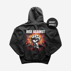 May include: Dark gray hoodie with the words "RISE AGAINST" above a graphic of a clenched fist breaking chains, set against a cityscape with red and orange hues. The brand name "MOSHPIT APPARELS" is printed on a black circle.