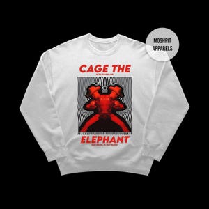 Può includere: Felpa bianca con la scritta "CAGE THE ELEPHANT" in rosso, sopra una grafica rossa e nera. La grafica è un'immagine stilizzata di due figure su uno sfondo a strisce bianche e nere. La felpa ha uno scollo tondo.