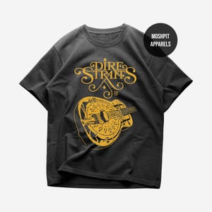 May include: Dark gray t-shirt featuring a distressed gold graphic of a mandolin and the band name "Dire Straits". The shirt also has the text "Moshpit Apparels" in a black circle. A classic music-themed tee.