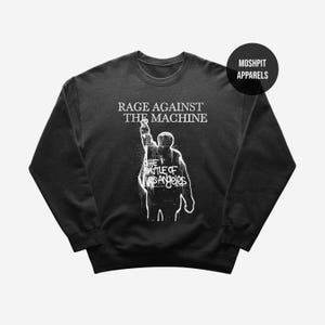 May include: Black crewneck sweatshirt featuring the text "RAGE AGAINST THE MACHINE" above a white graphic of a person raising their arm. Below the graphic is the text "THE BATTLE OF LOS ANGELES". The sweatshirt has a round logo that says "MOSHPIT APPARELS".