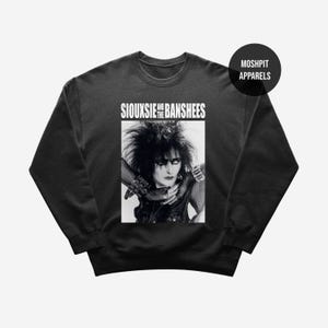 Pode incluir: Camisola cinza escuro com uma imagem a preto e branco de Siouxsie Sioux e o nome da banda "Siouxsie and the Banshees" em texto branco. A camisola tem gola redonda e mangas compridas. O nome da marca "Moshpit Apparels" está num círculo preto.