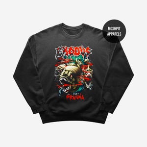 以下が含まれることがあります： ピラニアのグラフィックと、その上に赤い文字で「EXODUS」と書かれた濃いグレーのスウェットシャツ。「Piranha」の文字は赤で下に書かれています。スウェットシャツはクルーネックと長袖です。