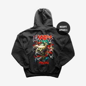 以下が含まれることがあります： ピラニアのグラフィックと赤い「Piranha」の文字が入った濃いグレーのパーカー。「Exodus」の文字は、赤と白でピラニアの上にあります。パーカーにはフードと「Moshpit Apparels」のロゴが付いています。