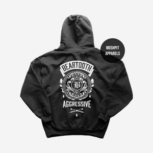 May include: A dark grey hooded sweatshirt featuring a white graphic on the back. The graphic includes the word "BEARTOOTH" above a circular design with the letter "B" and the word "AGGRESSIVE" below. A black circle with "MOSHPIT APPARELS" is on the upper right.