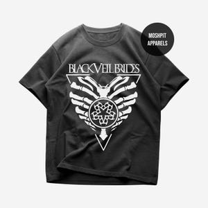 May include: Black t-shirt with a white graphic featuring the text "BLACK VEIL BRIDES" above a stylized design of a skeleton hand and a circular emblem. The shirt also has the text "MOSHPIT APPARELS" on a circular tag.