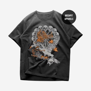 Puede incluir: Camiseta gris oscuro con un diseño gráfico detallado. El diseño incluye un pájaro estilizado con las alas extendidas, un pez y elementos ornamentados en naranja y blanco. La camiseta tiene el texto "Moshpit Apparels" en la parte superior derecha.