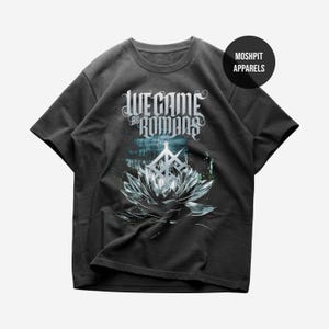 Könnte beinhalten: Dunkelgraues T-Shirt mit dem Text "We Came as Romans" in einer stilisierten Schriftart über einem detaillierten Lotusblumen-Design. Das Shirt hat auch ein Logo mit der Aufschrift "Moshpit Apparels".