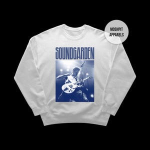 Puede incluir: Sudadera blanca con un gráfico azul de la banda Soundgarden, que muestra a un músico tocando la guitarra. El nombre de la banda está impreso encima de la imagen. La sudadera tiene cuello redondo y mangas largas.