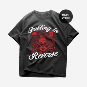 May include: Charcoal grey t-shirt with a red and white graphic design. The design includes a woman's face, skeletal hands, a skull, and the text "Falling in Reverse". The tee has a crew neck and short sleeves.