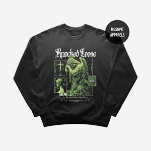Puede incluir: Sudadera negra de cuello redondo con un diseño gráfico. El diseño presenta una estatua de ángel, una cruz y las palabras "Knocked Loose" y "Blinding Faith" en blanco. El diseño también incluye las letras "K/L" en un recuadro verde.