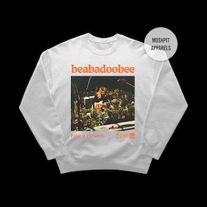 May include: White sweatshirt featuring the name "beabadoobee" in orange, above a photo of flowers and a person. The words "Fake It Flowers" are below the photo in orange. A white circle with "Moshpit Apparels" is in the upper right.