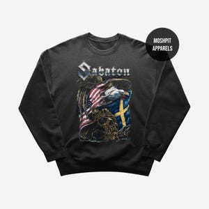 Könnte beinhalten: Dunkelgraues Sweatshirt mit einem grafischen Design, das das Wort "Sabaton", amerikanische und schwedische Flaggen, einen Totenkopf und einen Adler enthält. Das Sweatshirt hat einen Rundhalsausschnitt und lange Ärmel. Das Design ist auf der Vorderseite aufgedruckt.
