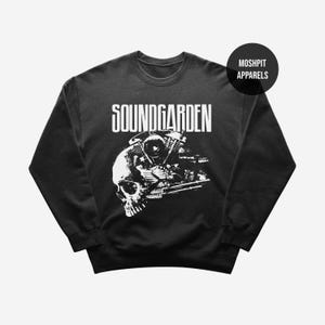 Puede incluir: Sudadera negra con un gráfico blanco de una calavera y un motor de motocicleta, con el nombre de la banda "SOUNDGARDEN" arriba. La sudadera también tiene un logotipo de "MOSHPIT APPARELS" en la parte superior derecha.