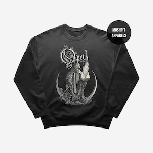 May include: Black crewneck sweatshirt featuring a detailed black and white illustration. The design includes the band name "Opeth" in an ornate font, a figure holding a book with flames, and a crescent moon. The brand name "Moshpit Apparels" is also visible.