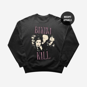 Może przedstawiać: Czarna bluza z okrągłym dekoltem z różową grafiką zespołu Bikini Kill. Grafika zawiera nazwę zespołu i stylizowany wizerunek członków zespołu. Bluza ma okrągły dekolt i długie rękawy.
