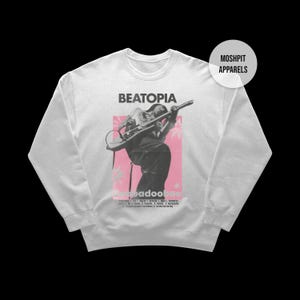 May include: White sweatshirt with a graphic of a person playing a guitar, set against a pink backdrop with the word "BEATOPIA" above. The design includes the text "Baddooloo" and other text at the bottom. The brand name "MOSHPIT APPARELS" is in a circle.