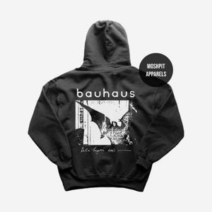 Può includere: Felpa con cappuccio grigio scuro con grafica bianca sul retro. La grafica presenta il nome della band "bauhaus" sopra un'immagine in bianco e nero e il testo "Bela Lugosi's dead". Un logo "Moshpit Apparels" è in alto a destra.