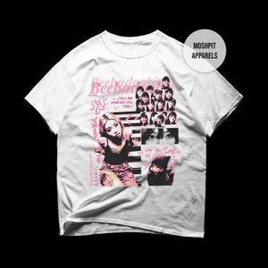 May include: White t-shirt featuring a pink and black graphic design. The design includes the text "Beeladoochie" and images of women. The shirt also has the text "Tell me how do you feel?" and the words "Moshpit Apparels".