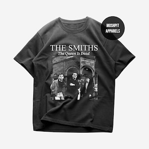 Op de afbeelding: Zwart T-shirt met de tekst "The Smiths The Queen Is Dead" boven een zwart-wit foto van de band. De afbeelding bevat de tekst "Salford Lads Club".