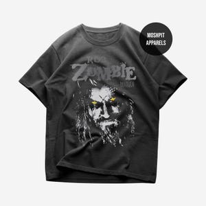 May include: Dark gray t-shirt featuring a graphic of Rob Zombie with the text "ROB ZOMBIE DRAGULA". The image shows a close-up of Rob Zombie's face with yellow eyes and a long beard. The shirt has a crew neck and short sleeves.