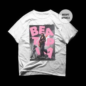 May include: White t-shirt with a graphic of a woman playing guitar on stage, overlaid with pink text reading "BEAT TOP IA". The shirt has a crew neck and short sleeves, with the "Moshpit Apparels" logo on the upper right.