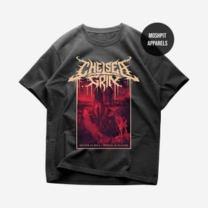 Puede incluir: Camiseta gris oscuro con un diseño gráfico. El diseño presenta el texto "THEISER GRIM" en una fuente estilizada sobre una ilustración roja y negra. La ilustración representa una escena con figuras y el texto "SUFFER IN HELL / SUFFER IN HEAVEN".