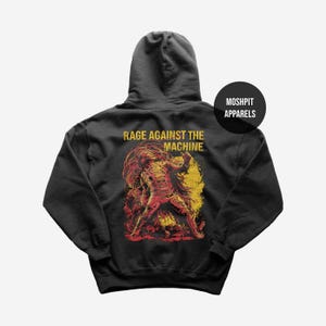 Puede incluir: Sudadera con capucha gris oscuro con un gráfico de una figura con llamas rojas y amarillas, con el texto amarillo "RAGE AGAINST THE MACHINE". La sudadera con capucha también tiene el texto "MOSHPIT APPARELS" en un círculo negro.