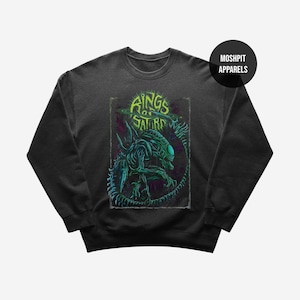 Op de afbeelding: Donkergrijze sweatshirt met een neon groen en blauw alien-ontwerp en de tekst "Rings of Saturn". Het ontwerp staat in een rechthoekige lijst. De merknaam "Moshpit Apparels" is op een zwarte cirkel gedrukt.