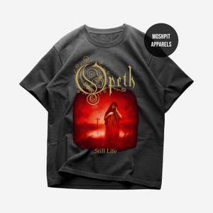 Puede incluir: Camiseta gris oscuro con el nombre de la banda "Opeth" en letras doradas sobre un gráfico rojo. El gráfico representa una figura con una túnica roja frente a una cruz, con las palabras "Still Life" debajo.