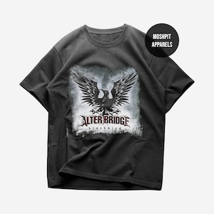 Op de afbeelding: Donkergrijs t-shirt met een zwarte adelaar design en de tekst "ALTER BRIDGE blackbird" in rood en wit. Het shirt heeft een ronde hals en korte mouwen. De merknaam "MOSHPIT APPARELS" staat in een zwarte cirkel.
