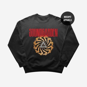 Puede incluir: Sudadera negra de cuello redondo con el nombre de la banda "SOUNDGARDEN" en letras rojas sobre un gráfico dorado y negro. El gráfico incluye un triángulo con texto y un diseño circular.