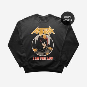 May include: Black crew neck sweatshirt with a graphic design. The design features the word "ANTHRAX" above an image of a helmeted figure, with the text "I AM THE LAW" below. The design is in yellow, red, and black.