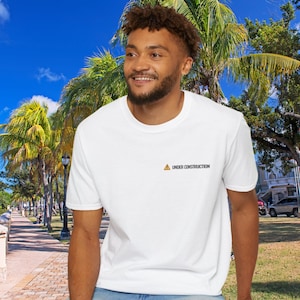 Könnte beinhalten: Weißes T-Shirt mit dem Text "UNDER CONSTRUCTION" in Schwarz. Das T-Shirt hat einen Rundhalsausschnitt und kurze Ärmel. Das Bild wurde im Freien mit Palmen und blauem Himmel im Hintergrund aufgenommen.