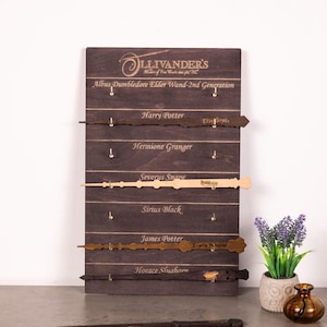 May include: A dark brown wooden display board featuring the Ollivander's logo and text for Albus Dumbledore, Harry Potter, Hermione Granger, Severus Snape, Sirius Black, James Potter, and Horace Slughorn, with four wands mounted on gold hooks.