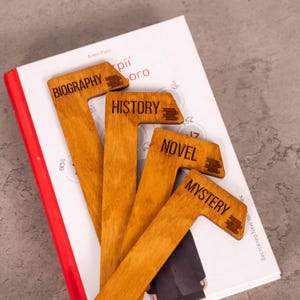 Può includere: Segnalibri in legno con le parole "Biography", "History", "Novel" e "Mystery" incise. I segnalibri sono disposti sopra un libro con copertina bianca e dorso rosso. I segnalibri sono di colore marrone chiaro.