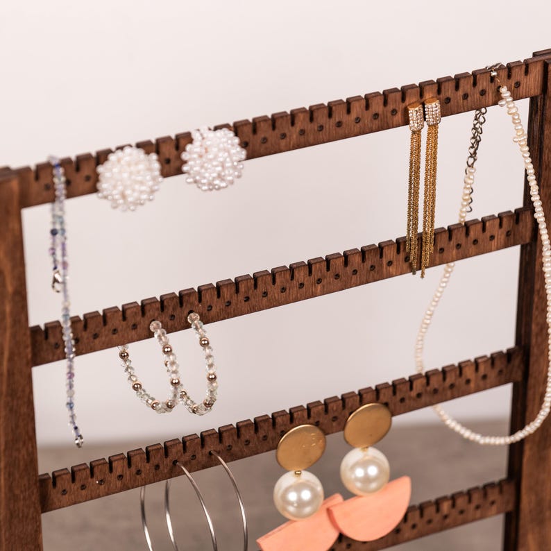 Puede incluir: Un expositor de joyas de madera marr&oacute;n oscuro con cuatro barras horizontales con bordes dentados para colgar accesorios. El soporte sostiene varios pendientes, incluyendo racimos de perlas, borlas de cadena dorada, aros de cuentas, aros plateados y colgantes color melocot&oacute;n.