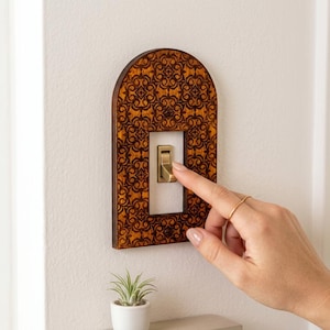 Pode incluir: Uma placa de interruptor de madeira em forma de arco com um padrão floral intrincado gravado a laser em castanho escuro. A placa está montada numa parede e rodeia um interruptor de alavanca retangular cor de latão. A madeira tem um acabamento castanho médio.