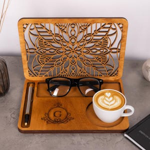 May include: A wooden desk organizer featuring an upright laser-cut mandala panel, a dedicated slot for a pen, a rectangular compartment holding black-rimmed eyeglasses, and a circular coaster holding a white ceramic cup of latte with foam art.